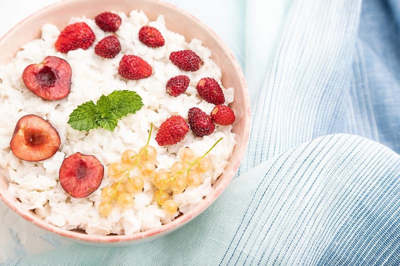 Rice flakes porridge with milk and strawberry in ceramic bowl on white concrete background and blue linen textile. Top view, flat lay, close up