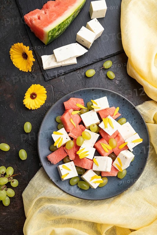 Vegetarian salad with watermelon, feta cheese, and grapes on blue ceramic plate on black concrete background and yellow linen textile. top view, close up, flat lay