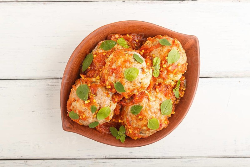 Pork meatballs with tomato sauce, oregano leaves, spices and herbs in clay bowl on a white wooden background. top view, flat lay, close up