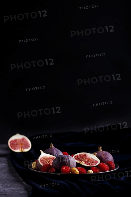 Fresh figs, strawberries and raspberries on blue ceramic plate on black concrete background and blue velvet textile. side view, copy space, selective focus, low key