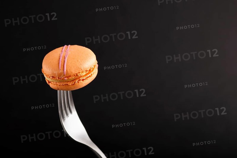 Single orange macaron or macaroon cake on a fork on black background. side view, close up, copy space