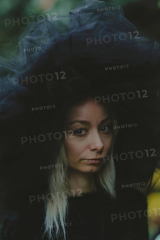 A woman wearing a tulle hat appears thoughtful, captured outdoors in natural lighting