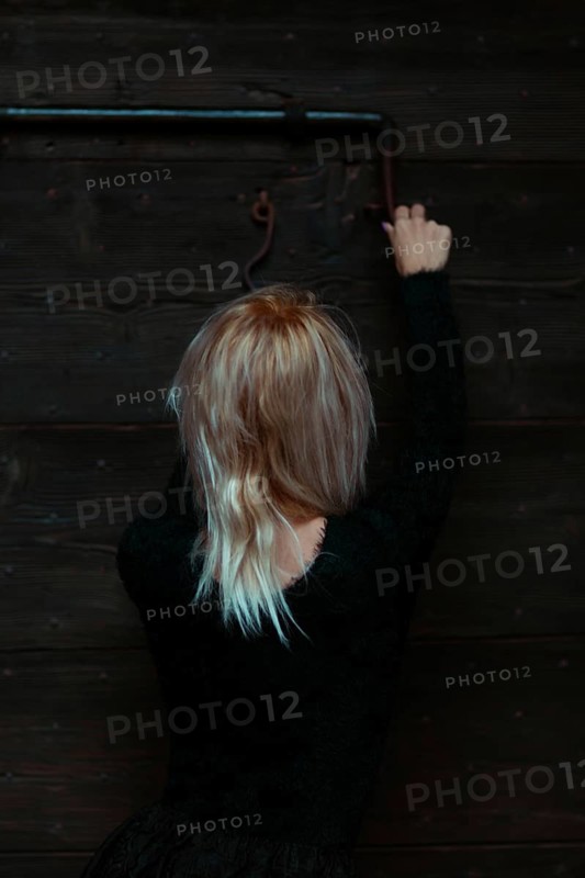 Back view of a blonde thin white woman dressed in black reaching up to a rusty antique metal lock on a wooden door