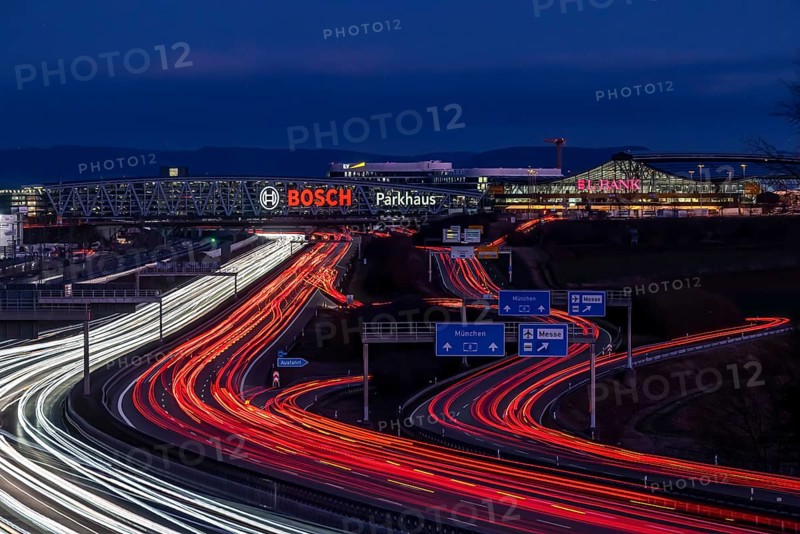 A8 motorway at Stuttgart Airport with Bosch multi-storey car park in the evening, cars create light trails. The 440 metre long construction offers space for 4200 vehicles. Bosch owns the naming rights, the stylised ignition anchor measures 12 metres in diameter. This makes it one of the largest neon signs in the world. Stuttgart, Baden-Wuerttemberg, Germany, Europe