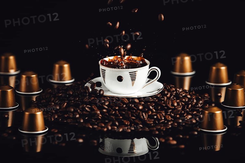 Coffee cup on coffee beans with falling beans and splashes, coffee capsules, reflection, black background