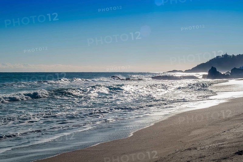 Sunlit waves crash against a rocky shore with glistening sea foam, in South Korea