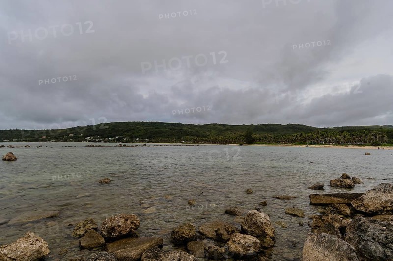 Seascape of rocky shoreline on a cloudy day with buildings on tree lined shore in the distance in Guam