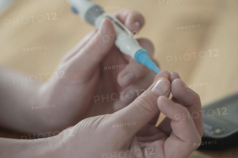 On the left a child's hand holding a lancet, lancing device, and pricking itself to determine the blood glucose level, wooden table as background, measuring device device blurred on the right, blood glucose measurement, diabetes treatment, glucose measurement, Ruhr area, Germany, Europe