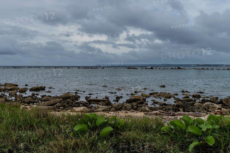 Seascape of rocky shoreline on a cloudy day with buildings on tree lined shore in the distance in Guam