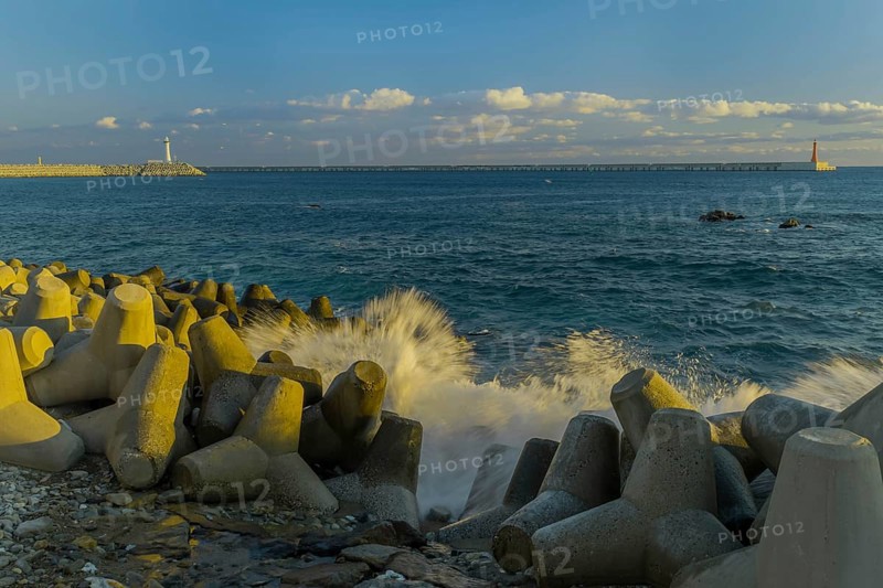 Waves crashing against tetrapods laying on ocean shoreline with two lighthouses on piers on horizon under partly cloudy morning sky, in South Korea