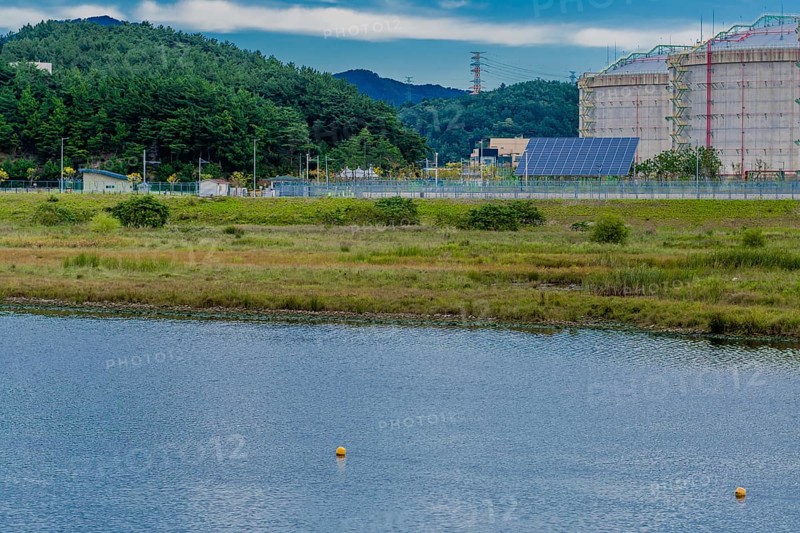 Industrial storage tanks beside a calm water body dotted with buoys, set against a mountain backdrop, in South Korea