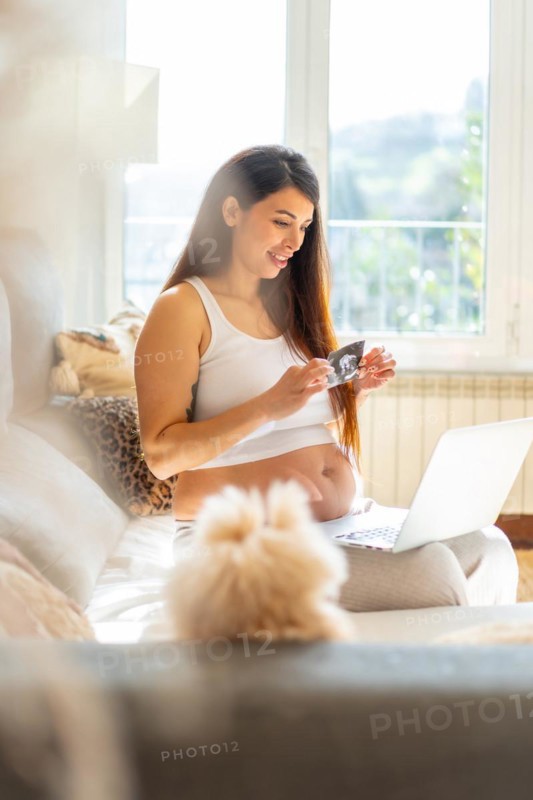 Vertical photo of a breauty adult pregnant woman showing baby ultrasound in video call using laptop sitting on the sofa at home