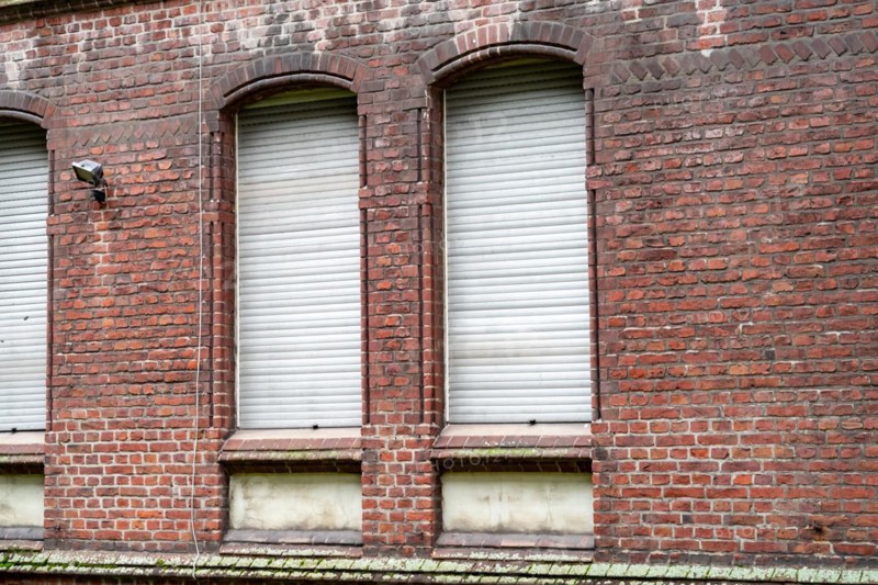 Three evenly spaced windows with closed white shutters in a red brick wall