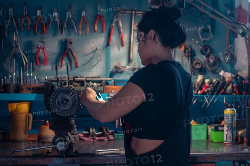 Skilled craftswoman repairing a motorcycle engine part in a workshop with a teal wall, a complete tool panel in background with bokeh effect, traditional male jobs by Mixed-race latino woman