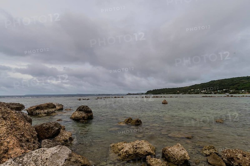 Seascape of rocky shoreline on a cloudy day with buildings on tree lined shore in the distance in Guam