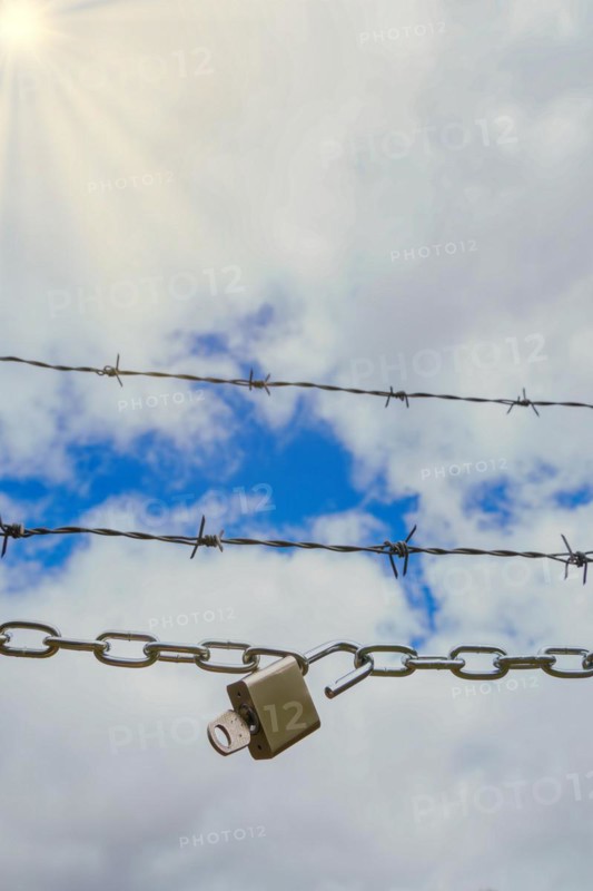 Chain with padlock open with key next to a barbed wire with a blue cloudy sky with sun rays, concept of freedom