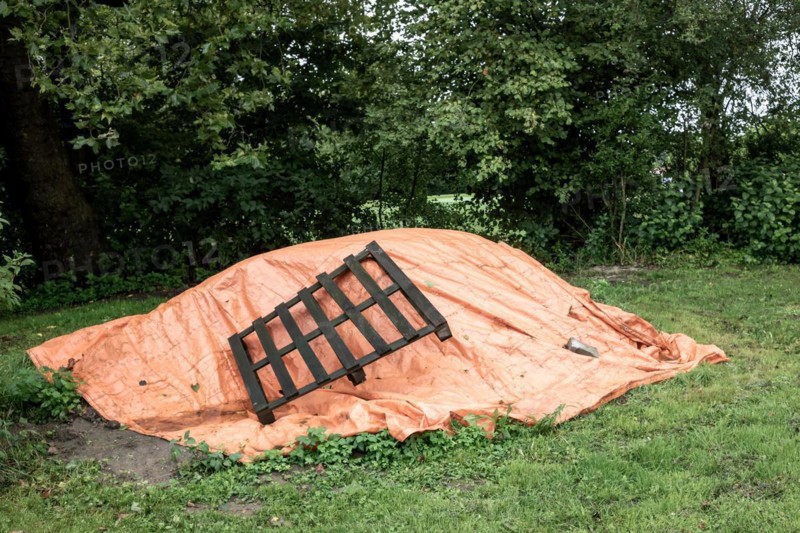 An orange tarpaulin covers something on the ground next to a wooden pallet and bushes