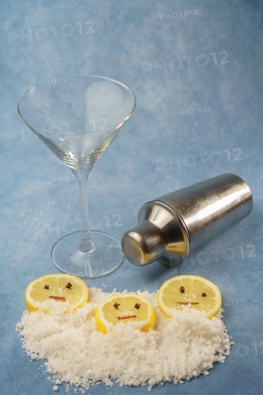 Lemon slices with cloves and cinnamon forming a smiley face on a pile of salt next to a cocktail shaker and a crystal glass with blue background with copy space