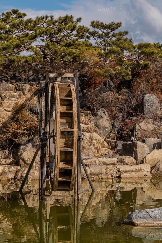 Close-up of a water wheel with its reflection in a pond, set against a rocky backdrop, in South Korea