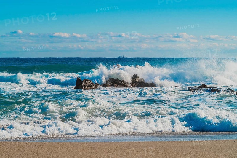 Crashing waves create spray around rocks on a sunlit beach with distant horizon, in South Korea