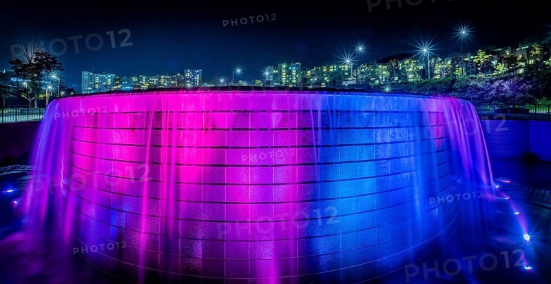 Night scene of waterfall lit up with blue and red lights with the lights of buildings of a town in South Korea in the background in Sejeong, South Korea, Asia