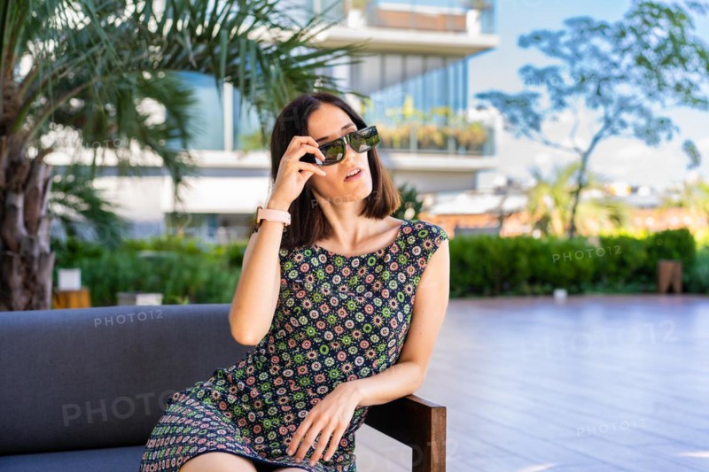 A beautiful woman is sitting on a couch outdoors while taking off her sunglasses