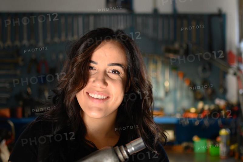 Joyous woman mechanic laughing in a workshop environment, a complete tool panel in background with bokeh effect, traditional male jobs by Mixed-race latino female