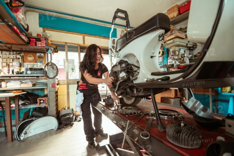 A woman mechanic works on a vintage scooter in a cluttered workshop, wearing black overall and shirt, latino female in traditional masculine jobs concept, feminine power in real life