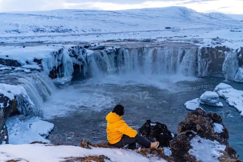 Adventurous woman in winter in Iceland sitting looking at frozen Godafoss waterfall
