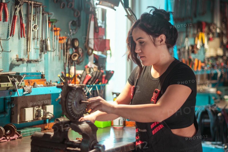 A woman is attentively working on a mechanical piece at a tidy workshop bench, a complete tool panel in background with bokeh effect, traditional male jobs by Mixed-race latino woman