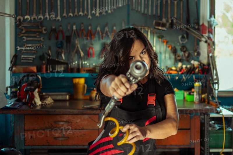 Intense sexy woman mechanic aiming a tool at the camera in a workshop, a complete tool panel in background with bokeh effect, traditional male jobs by Mixed-race latino woman