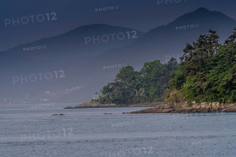 Landscape of shoreline with lush green trees and small mountains shrouded in fog and mist in the background in Namhae, South Korea, Asia