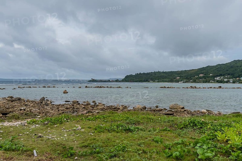 Seascape of rocky shoreline on a cloudy day with buildings on tree lined shore in the distance in Guam