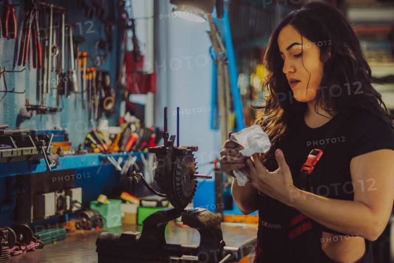 A female mechanic wearing overall in a workshop is carefully cleaning motorcycle parts, a complete tool panel in background with bokeh effect, traditional male jobs by Mixed-race latino woman