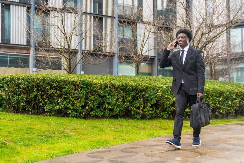 African young businessman commuting while talking to the mobile outdoors along a public urban park