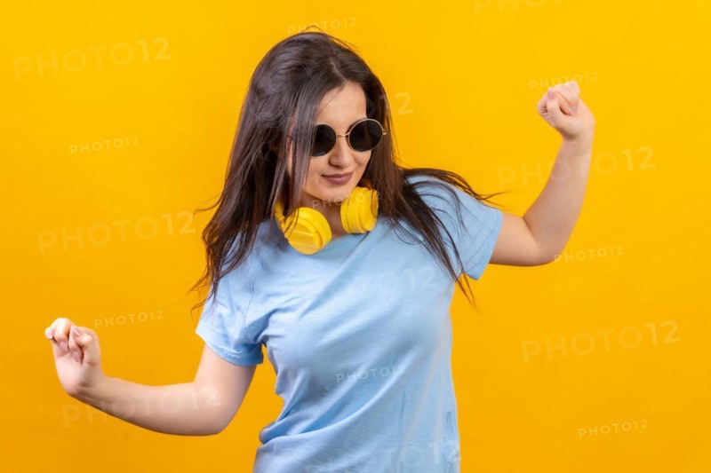Studio portrait with yellow background of happy woman dancing wearing sunglasses and headphones on the neck