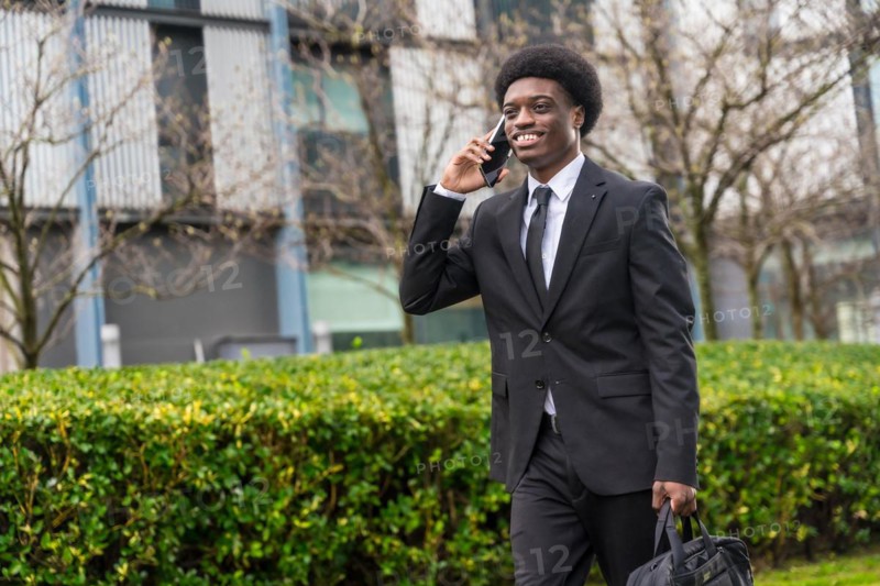 Three quarter length portrait of a cool smiling african businessman walking along the street using phone