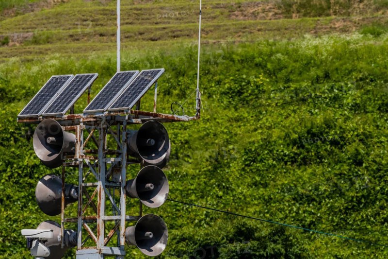 Communication tower consisting of loud speakers, solar panel and closed circuit camera with tree covered mountain in background in South Korea