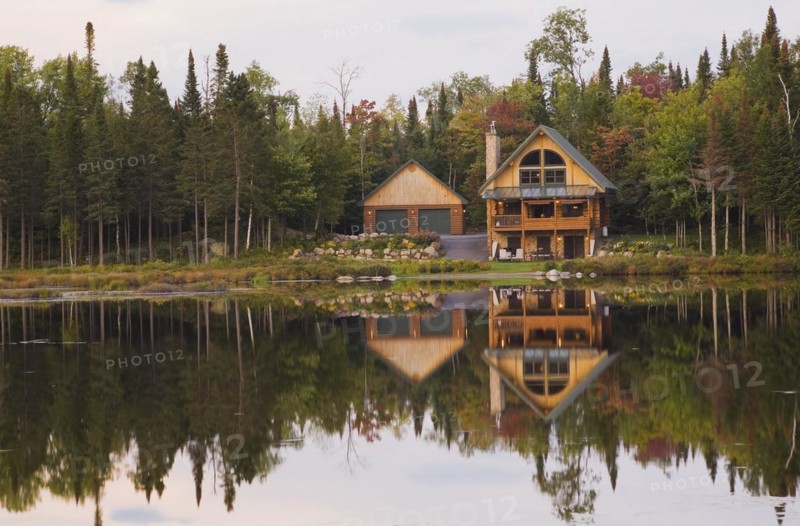 Garage and handcrafted two story spruce log cabin home with fieldstone chimney and green sheet metal roof on edge of lake in autumn, Quebec, Canada, North America