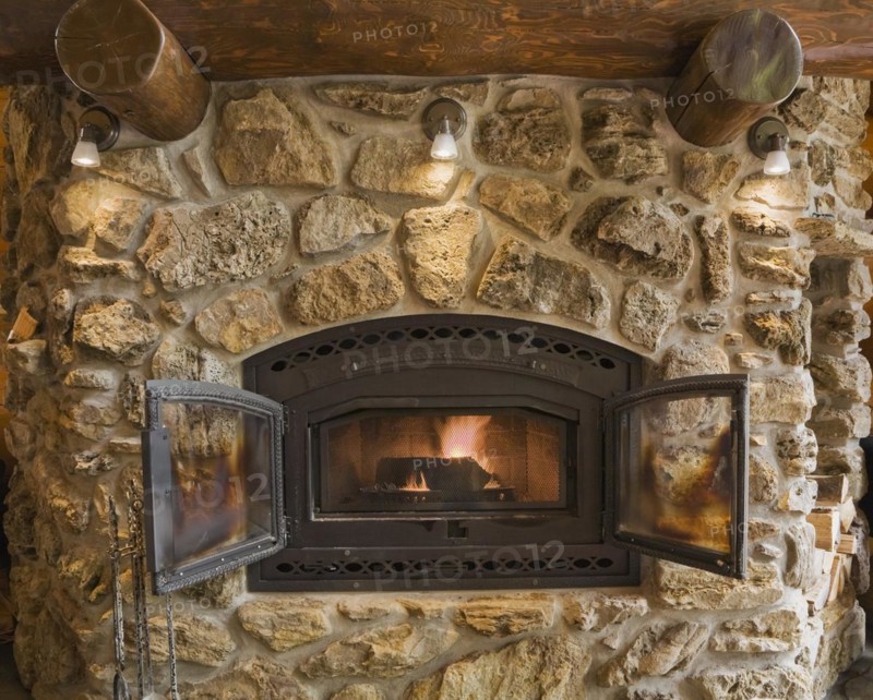 Lit fieldstone and porous rock fireplace in living room inside handcrafted red cedar log cabin home, Quebec, Canada, North America