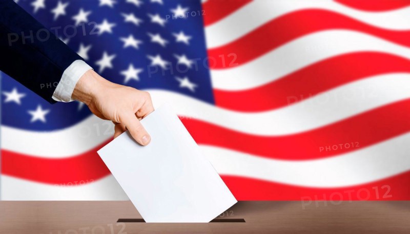 USA presidential elections. Hand holding ballot in voting ballot box with USA flag in background