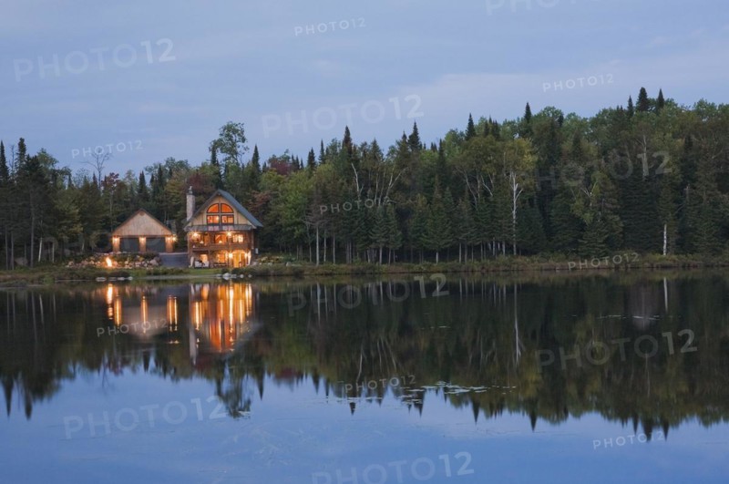 Illuminated two car garage and handcrafted two story spruce log cabin home with fieldstone chimney and green sheet metal roof on edge of lake at dusk in late summer, Quebec, Canada, North America