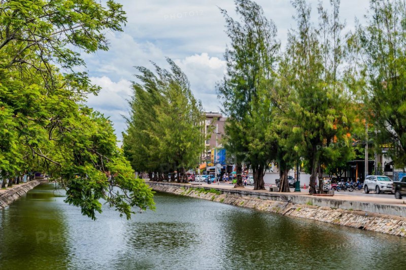 Cloudy sky reflecting on canal water, with a vibrant urban tree-lined waterfront, in Chiang Mai, Thailand, Asia