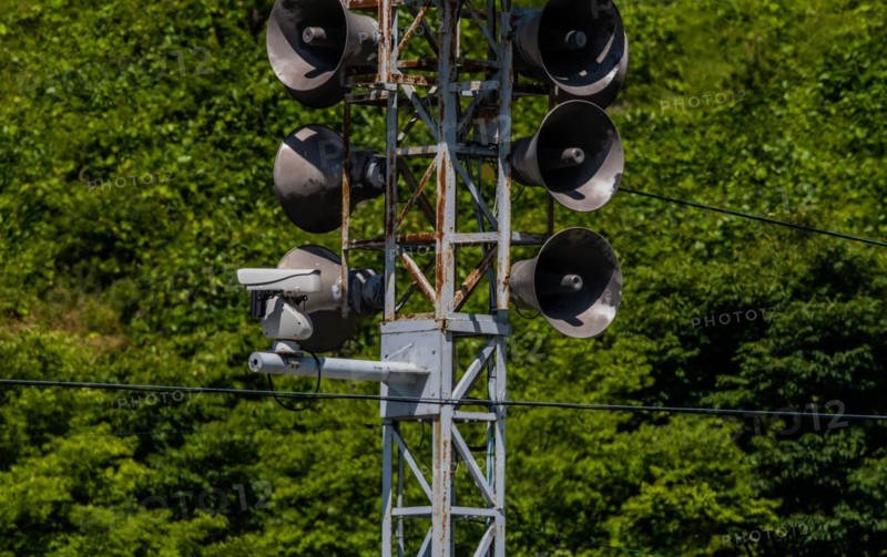 Communication tower consisting of loud speakers, solar panel and closed circuit camera with tree covered mountain in background in South Korea