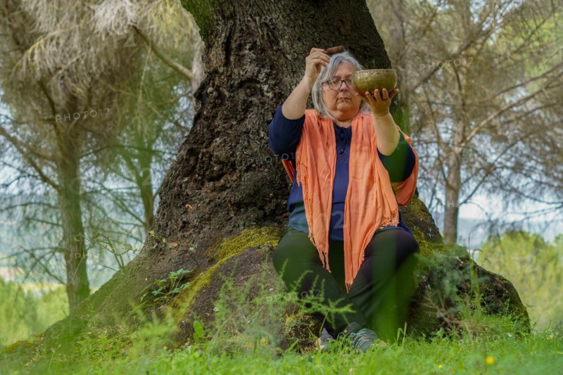 Mature woman with white hair meditating in the forest with her eyes closed and a Tibetan singing bowl ringing