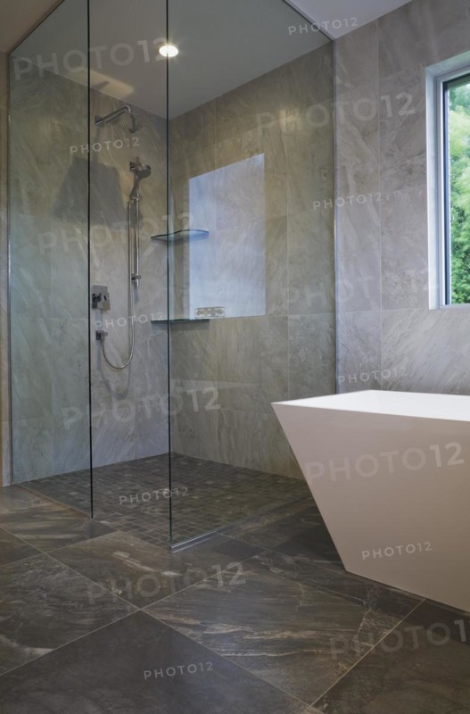 Clear glass shower stall and white freestanding vessel shaped bathtub in bathroom with grey ceramic tile floor and walls on ground floor inside modern cubist style home, Quebec, Canada, North America