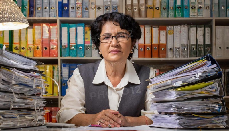 A woman sits behind a desk full of folders and piles of paper in the office, symbolising bureaucracy, AI generated, AI generated