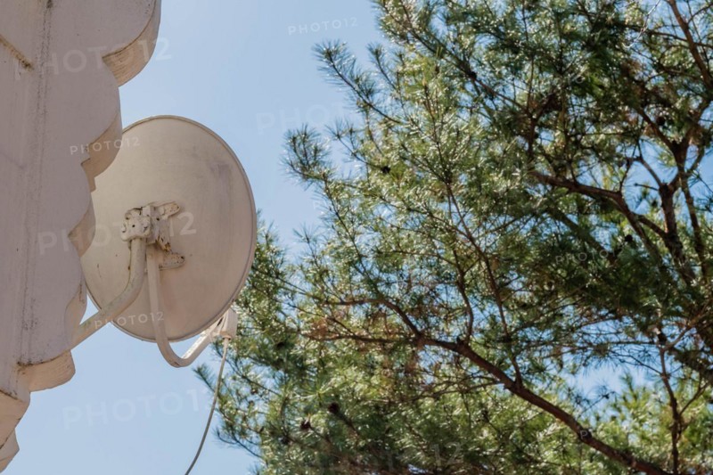 A white satellite dish mounted on a wall with a tree and blue sky in the background, in South Korea