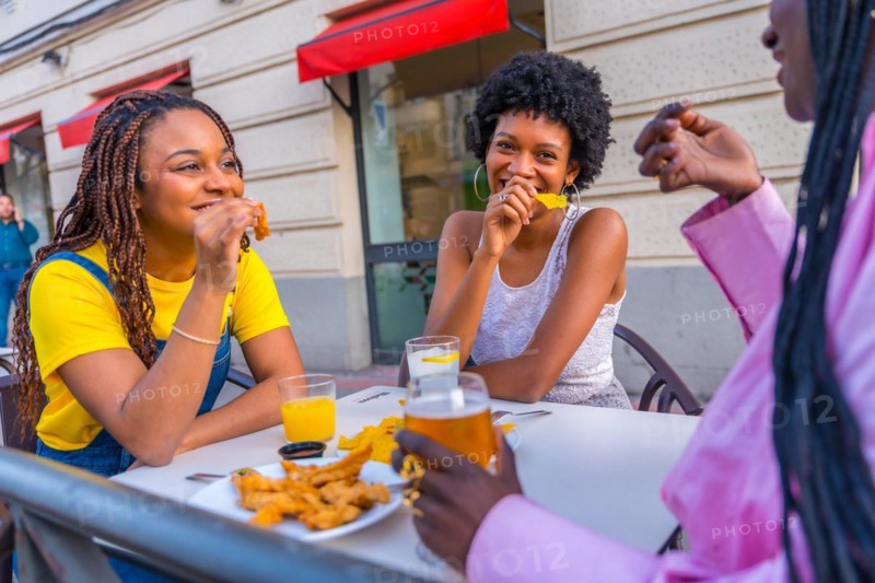Three female young african friends eating fast food with beer in an outdoor restaurant