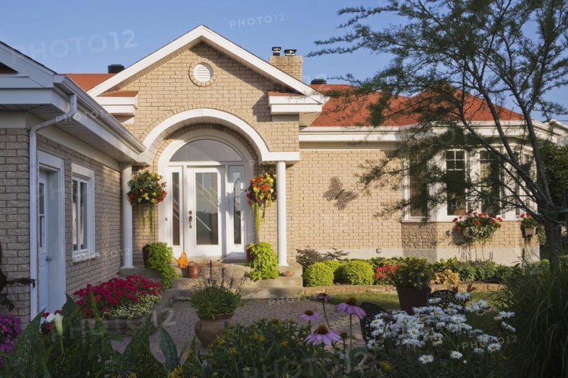 Tan brick home with white trim and landscaped front yard with raised border with red Pelargonium, Geranium flowers, Echinacea purpurea, Coneflowers and white Leucanthemum, Oxeye daisies in summer, Quebec, Canada, North America